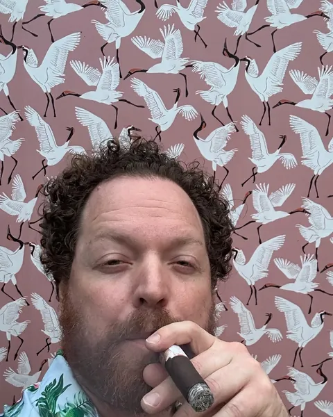 Dave Menzo with a cigar in a Hawaiian shirt against a paper-crane wallpaper